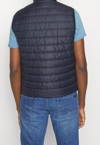 Marineblaues Steppwesten ohne Ärmel mit horizontalen Nähten und hohem Kragen, getragen über einem blauen T-Shirt. Kombiniert mit blauen Jeans.