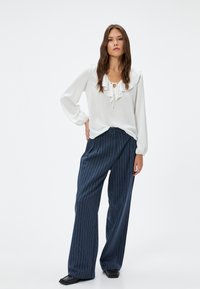 Witte blouse met een gerimpelde halslijn en strikdetail, gecombineerd met marineblauwe gestreepte wijde broek. De outfit heeft gladde texturen en ontspannen vormen.