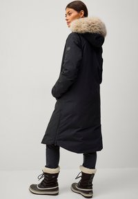 Zwarte lange parka met een nepbont capuchon, ritsventilatie aan de zijkanten en twee zijzakken. Draag het over donkere jeans en zwarte winterlaarzen met bontafwerking.