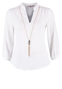 Blouse blanche à manches longues avec encolure en V, épaules froncées, et un long collier chaîne doré avec un pendentif tassel noir et doré.