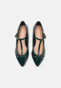 Green patent leather shoes with a T-strap design, pointed toes, and a decorative bow at the front. Features a beige insole with branding.