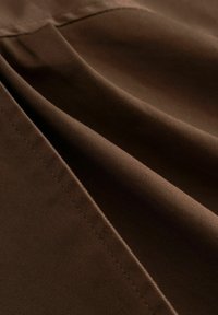 Hnedá tkanina s hladkou textúrou, obsahujúca jemné diagonálne čiary a záhyby, ktoré ukazujú strednú hmotnosť a matný povrch.