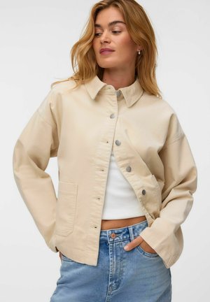 Kvinde med blondt hår iført en beige knapjakke over en hvid crop top og blå jeans, med den ene hånd i lommen.