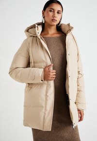 Veste rembourrée beige avec une capuche, dotée d'une fermeture éclair, de boutons-pression et de poches latérales. Portée sur une robe brune à côtes.
