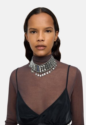 Srebrny naszyjnik w formie ozdobnej, składający się z geometrycznych elementów metalowych, zestawiony z czarną zwiewną bluzką i elegancką czarną bielizną.