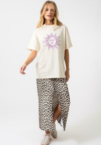 T-shirt en coton couleur crème avec un graphique de soleil violet, associé à une jupe maxi à imprimé léopard avec une fente sur le côté et des baskets beige.