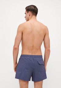 Blå og hvite stripete svømshorts med elastisk liv og sidelommer, vist fra baksiden. Stoffet ser lett og teksturert ut.