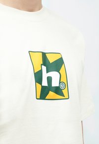 Κρεμ T-shirt με γραφικό πράσινου και κίτρινου αστεριού που έχει μέσα ένα λευκό πεζό «h» και ένα κυκλικό λογότυπο «HUF». Μαλακό ύφασμα.