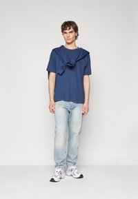 Camiseta de algodón azul marino llevada con un tejido similar atado en un nudo sobre los hombros, combinada con jeans de un azul claro y zapatillas blancas.