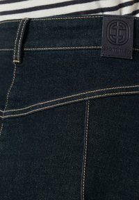 Tmavé džíny mají hladkou texturu, zlaté prošití a značkovou nášivku na pásu. Je viditelný klasický pětikapsový design.