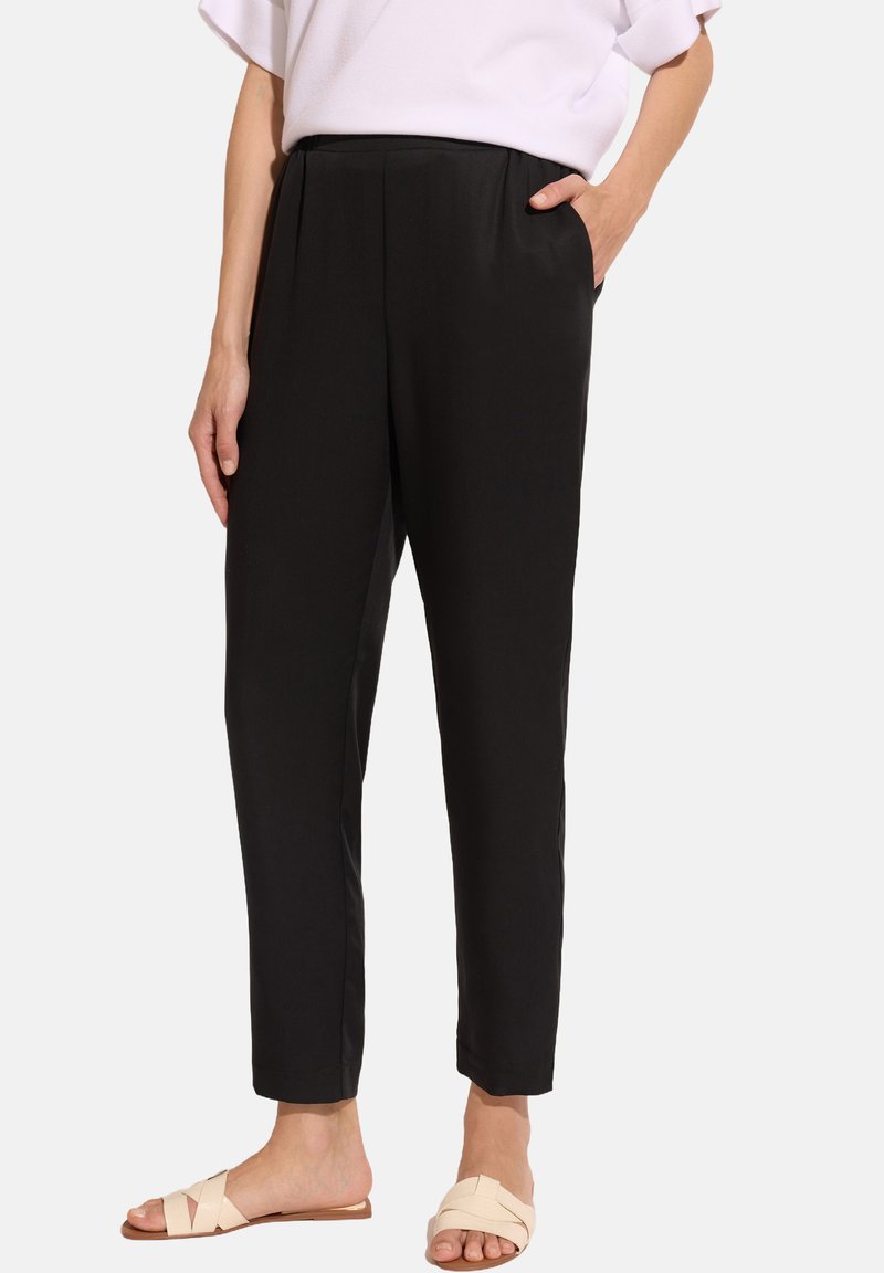 Czarne, tapered spodnie z elastycznym pasem i bocznymi kieszeniami, wykonane z gładkiego materiału. Ubrane z jasnymi sandałami.
