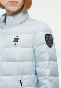 Νεαρό άτομο που φοράει ανοιχτό γαλάζιο μπουφάν puffer με ετικέτα "Blauer U.S.A." στο στήθος και έμβλημα-ασπίδα στον δεξί ώμο.