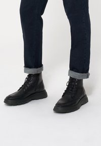 Czarne skórzane botki z sznurówkami, gumową podeszwą, zaokrąglonym noskiem i wyściełanym kołnierzem. Noszone z podwiniętymi ciemnymi dżinsami.