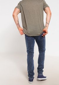 Camiseta de manga corta gris con un corte holgado, combinada con vaqueros azul oscuro de corte ajustado y zapatillas azules. Tatuajes visibles en los brazos.