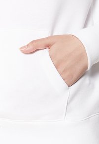 Sudadera de algodón blanca con un bolsillo frontal, mostrando una mano descansando dentro. La tela tiene una textura suave y las costuras están bien terminadas.