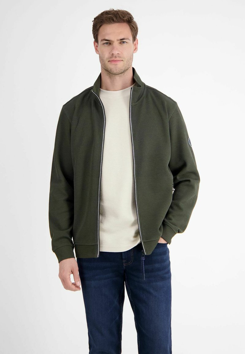 Dunkelgrüner Reißverschlussjacke mit Stehkragen, strukturiertem Strickmuster und kontrastierendem hellgrauen Reißverschluss, kombiniert mit einem hellbeigen T-Shirt und Jeans.