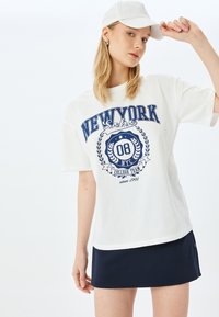 Hvid bomulds t-shirt med blå "NEW YORK Soho" grafik, korte ærmer, løst fit, parret med en marineblå nederdel, model iført en hvid cap.