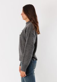 Donna con lunghi capelli castani, che indossa un maglione lavorato a maglia grigio e jeans blu, in piedi di profilo contro uno sfondo bianco semplice.