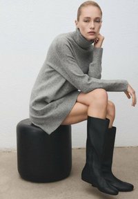 Sivi pulover haljina s rebrastom teksturom uparen s crnim čizmama do koljena. Model sjedi na okrugloj crnoj stolici. Minimalistička pozadina.