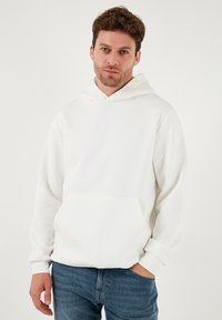 Hvid pullover-hoodie lavet af blødt stof, med en forlomme foran, ribstrikkede manchetter og en afslappet pasform med en rummelig hætte.