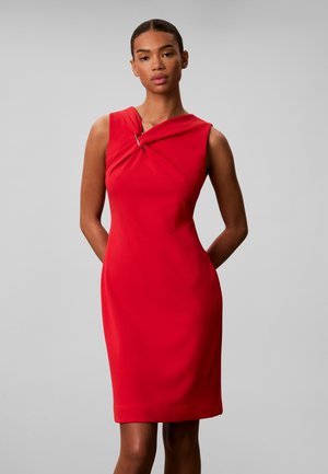 Femme avec les cheveux tirés en arrière portant une robe rouge sans manches avec un décolleté asymétrique et un détail d'anneau métallique.