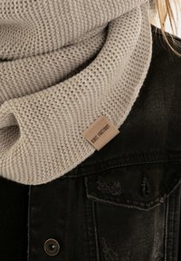 Lichtgrijze gebreide sjaal met een gestructureerd patroon, voorzien van een bruine leren label met de tekst "Knit Factory," over een zwart spijkerjacket gedrapeerd.