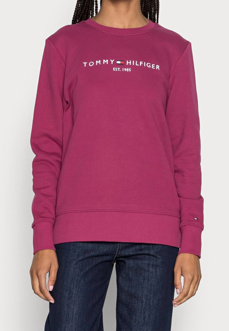Personne portant un sweat-shirt à manches longues Tommy Hilfiger rose foncé avec logo et un jean bleu foncé, debout les bras détendus le long du corps.