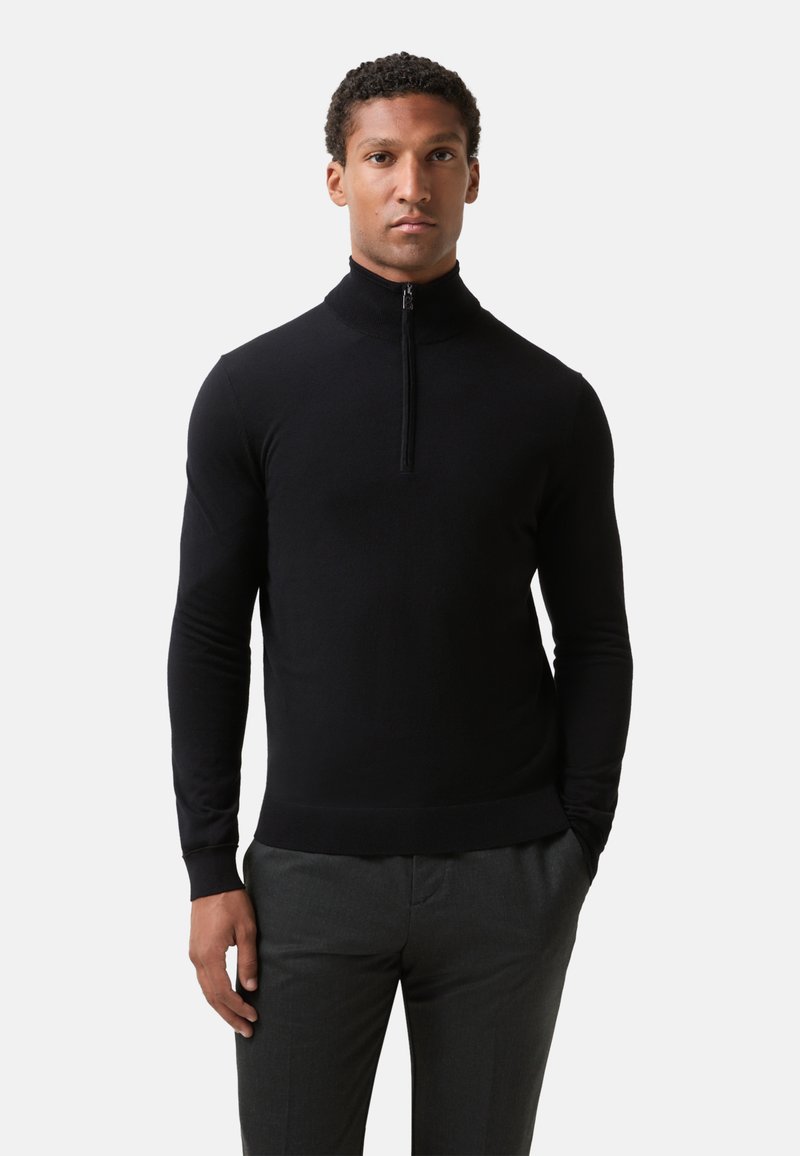 Schwarzer Pullover mit Reißverschluss am Hals und langen Ärmeln, hergestellt aus glattem Stoff. Verfügt über eine taillierte Passform sowie gerippte Bündchen und Saum. Kombiniert mit grauen Hosen.