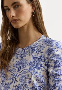 Top floreale blu con scollo rotondo e maniche lunghe, realizzato in un tessuto morbido con uno sfondo beige chiaro.