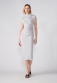 Sivá priliehavá šaty vyrobené z jemnej látky, s elegantným drapovaným výstrihom a dĺžkou po kolená. Model nosí čierne sandále na podpätku.
