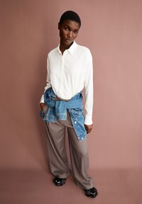 Camicia bianca con bottoni, giacca di denim legata in vita, pantaloni beige a gamba larga e scarpe nere con frange su uno sfondo rosa.