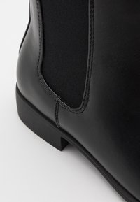 Czarne skórzane botki sięgające kostki z elastycznymi bocznymi panelami, płaską podeszwą i przeszyciami. Gładka faktura o matowym wykończeniu.