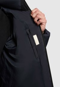 Tengerészkék zakó belseje sima textúrával, cipzáras zsebbel és rögzített címkével, valamint egy kontrasztos sötét panel a külső részen.