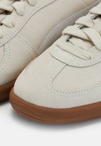 Gros plan sur des baskets en daim couleur crème avec des semelles en caoutchouc marron texturées et des lacets assortis couleur crème dans un style décontracté.