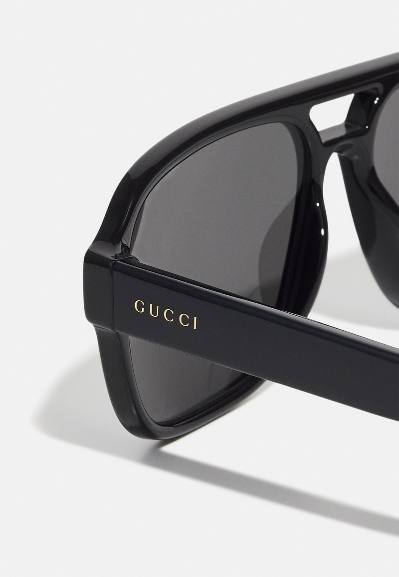 Lunettes de soleil Gucci avec des montures en plastique noir, des verres sombres et un détail de logo doré sur la branche. Elles présentent une forme rectangulaire moderne.