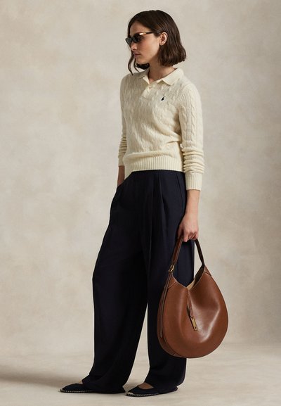 Vrouw draagt een crèmekleurige kabelgebreide trui, zwarte broek met brede pijpen, zwarte zonnebril, houdt een grote bruine leren handtas vast, staat tegen een neutrale achtergrond.