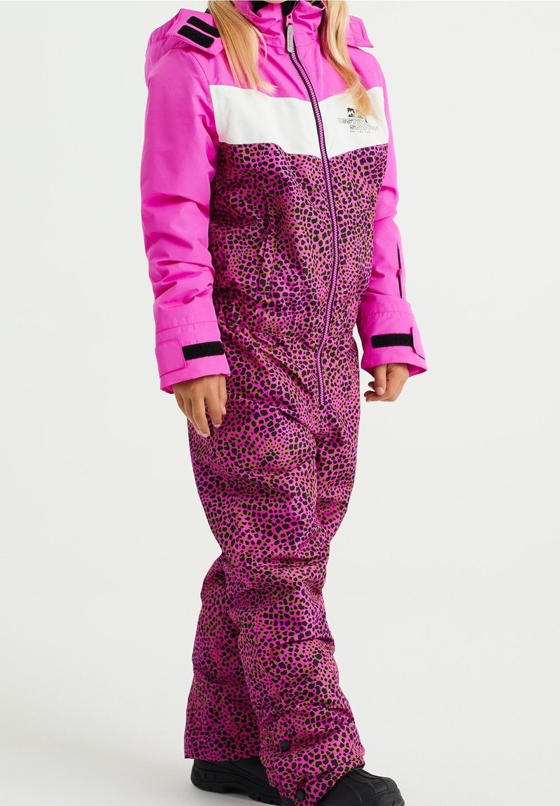 Barn klädd i en ljusrosa och leopardmönstrad snowsuit med svarta snöskor, stående mot en enkel vit bakgrund.