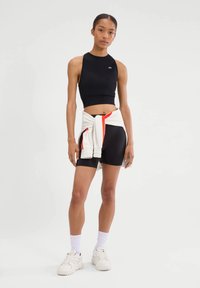 Zwart crop atletisch topje met een hoge hals, gecombineerd met zwarte korte broek. Een wit sweatshirt met rode accenten is om de taille gestrikt. Witte sneakers.