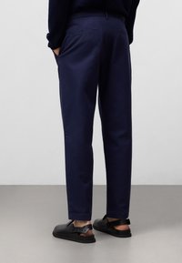 Navyblauwe broek in een slim fit ontwerp, gemaakt van soepele stof. Bevat twee achterzakken en heeft een net afgewerkte zoom. Draagt met zwarte sandalen.