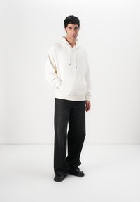 Weißer Kapuzenpulli mit Kordelzug und einer Fronttasche, kombiniert mit dunklen, weit geschnittenen Jeans und schwarzen Schuhen. Glatte Textur, lockere Passform.