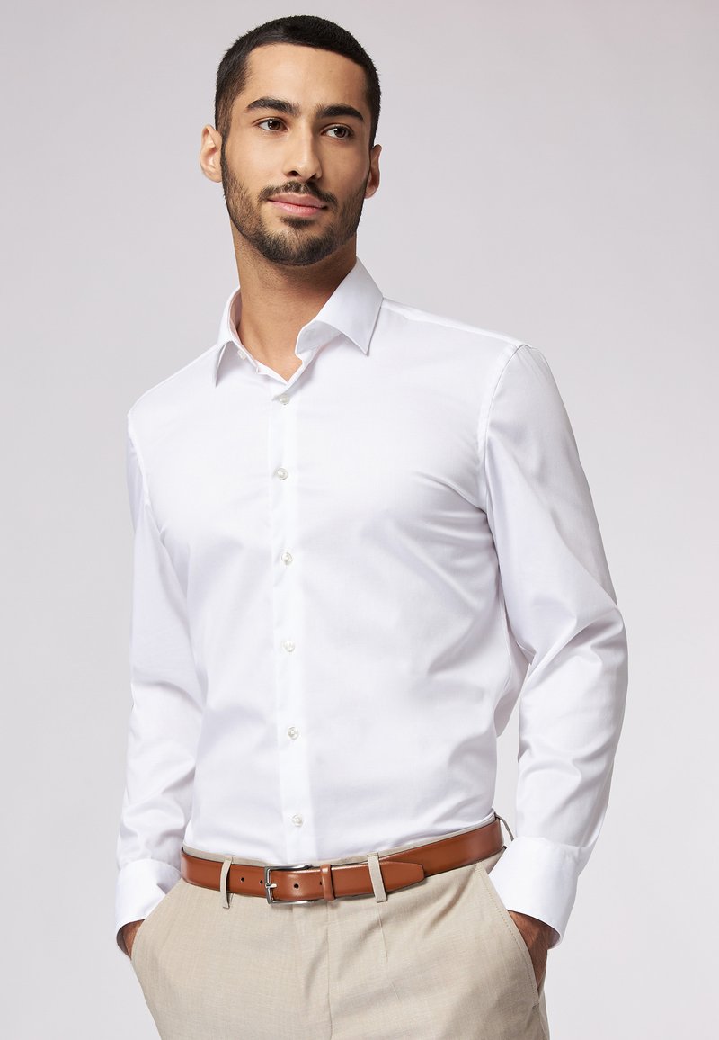 Weißes Langarmhemd mit spitzem Kragen, Knopfverschluss und glatter Textur, getragen mit hellfarbenen Hosen und einem braunen Gürtel.