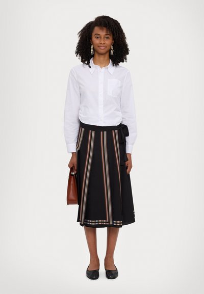 Camisa blanca de botones con un bolsillo, combinada con una falda negra con rayas verticales de colores y cintura anudada. Bolso marrón y bailarinas negras.