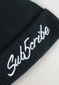 Beanie nera a maglia con texture a coste e testo ricamato bianco che recita "Sub5cribe." Finitura liscia senza ulteriori motivi o accenti.