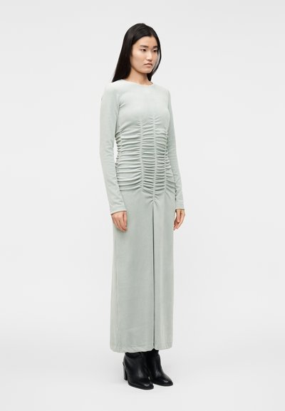 Obleka mint zelene barve z dolgimi rokavi in naborki vzdolž sprednjega dela in stranskih šivov, izdelana iz mehke tkanine. Stilizirana s črnimi škornji z visokimi petami.