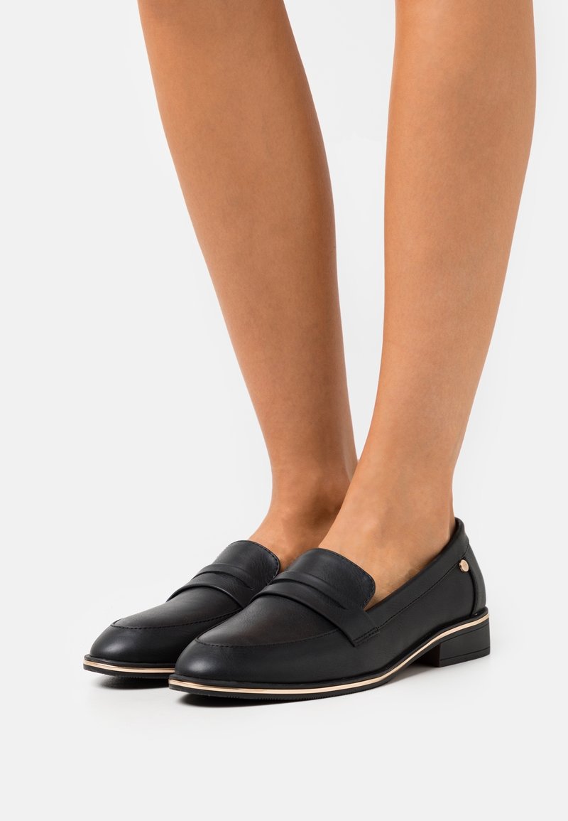Jambes portant des mocassins en cuir noir avec un petit talon et un bord beige, présentés sur un fond uni.