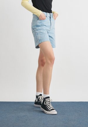 Osoba v svetlomodrých riflových šortkách, čiernych vysokých teniskách s bielymi šnúrkami, bielych ponožkách, čiernom tričku a žltých dlhých rukávoch, stojaca.