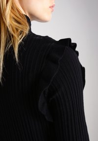Svart ribbad tröja med volangdetaljer vid axlarna. Slät yta, figursydd design, hög hals och diskret lyster.