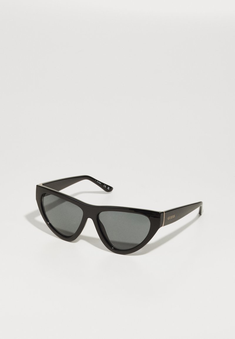 Ochelari de soare negri cu lentile întunecate, cu ramă din plastic lucios și un design ascuțit, având logo-ul auriu "GUESS" pe tempii.