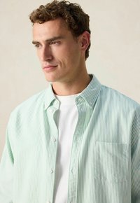 Jonge man met krullend haar, gekleed in een lichtgroen en wit gestreept overhemd over een wit T-shirt, kijkt naar de zijkant tegen een neutrale achtergrond.