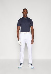 Camisa polo azul-marinho, calças brancas, cinto branco e sapatos de golfe brancos com detalhes azuis; segurando uma luva de golfe, em pé contra um fundo neutro.